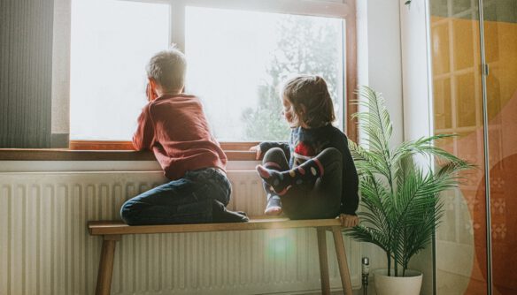 Kisgyerekek ülnek egy fapadon egy hosszú radiátor mellett az ablak előtt.
