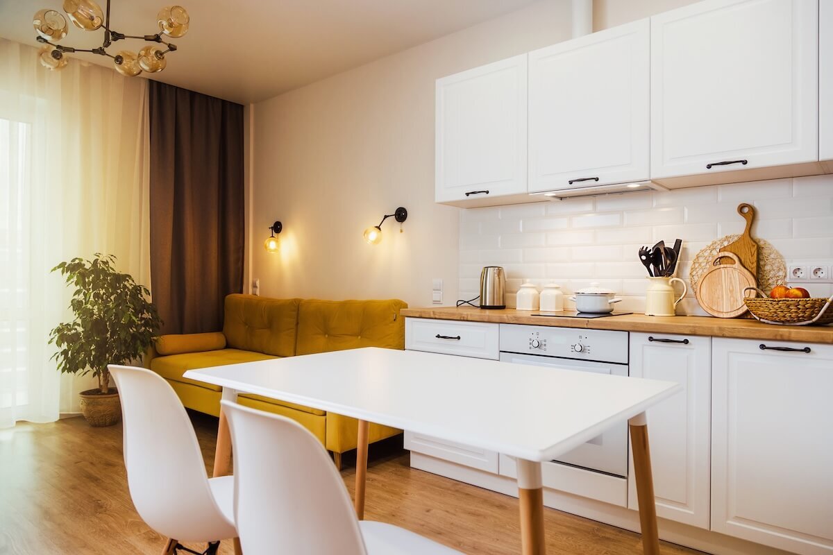 A képen egy modern, világos kis lakás nappali-konyha része látható, fehér bútorokkal és fa felületekkel berendezve. A tér otthonos hangulatát a sárga kanapé, meleg fényű lámpák és természetes díszítőelemek, például a növények és gyümölcskosár fokozzák.