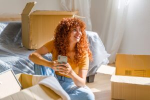 Fiatal nő költözés közben egy új otthonban, mosolyogva ül a földön okostelefonnal a kezében, körülötte nyitott kartondobozok.