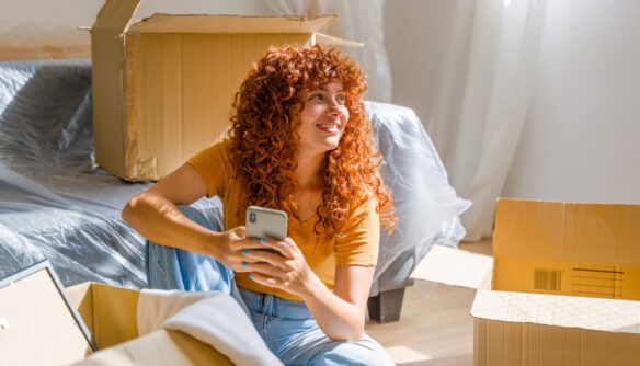 Fiatal nő költözés közben egy új otthonban, mosolyogva ül a földön okostelefonnal a kezében, körülötte nyitott kartondobozok.