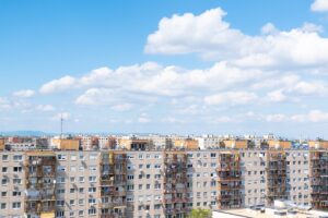 Panellakásokból álló lakótelep madártávlatból, tiszta kék ég és gomolyfelhők alatt. A képen többemeletes, erkélyes épületek sorakoznak, háttérben hegyek és városi panoráma sejlik fel.