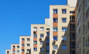 Panelház homlokzata kék ég alatt, több emeletes lakóépület ablakokkal és klímaberendezésekkel.