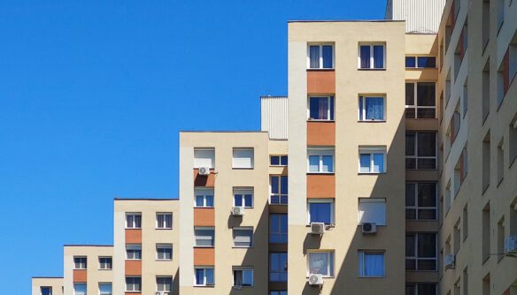 Panelház homlokzata kék ég alatt, több emeletes lakóépület ablakokkal és klímaberendezésekkel.