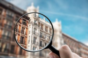 Kézben tartott nagyító fókuszál egy városi társasház homlokzatára, kiemelve a részleteket az elmosódott háttérből.