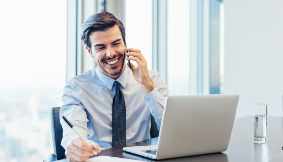 Elegánsan öltözött ingatlanközvetítő mosolyogva telefonál, előtte az asztalon laptop és papírok, mögötte az iroda hatalmas ablakai.
