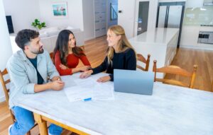 Fiatal pár ingatlanügynökkel tárgyal egy modern konyhában, az asztalon szerződés és laptop.