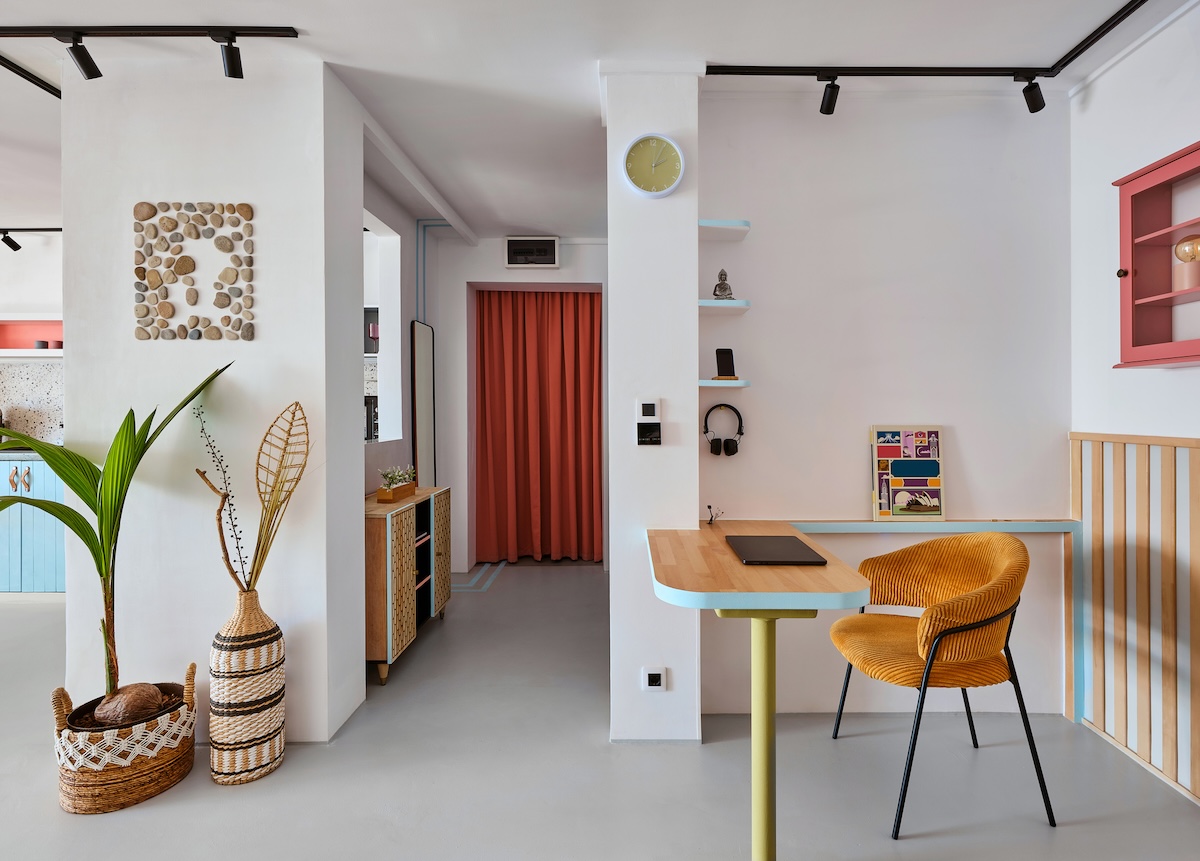 Világos, modern garzonlakás enteriőrje természetes anyagokkal és pasztellszínekkel. A helyiségben fa asztal kék éllel, sárga kárpitozott szék, polcokon dekorációk és fejhallgató, valamint terrakotta színű függöny látható. A sarokban zöld növények és fonott kosarak díszítik a teret, a falon kavicsképek és egy sárga falióra egészíti ki a harmonikus, otthonos hangulatot.
