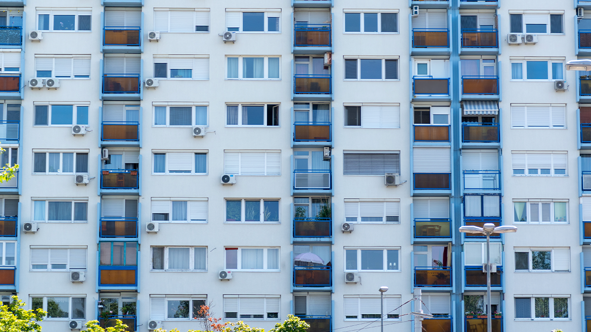 Panelház homlokzata kék erkélykorlátokkal és barna üvegfelületekkel. Az épület több emeletes, a lakások erkélyein különböző növények és napernyők láthatók. A falakon számos légkondicionáló berendezés található, az előtérben pedig néhány fa és utcai lámpa.