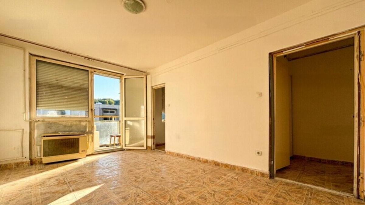 Üres, felújítandó lakás világos szobája, amelyben csempepadló, fehér falak és nagy ablakok láthatók. Az egyik falon régi gázkonvektor található, az ablakon redőny és egy ajtó vezet az erkélyre. A szoba mellett egy kisebb helyiség bejárata is látható, a tér napfényben úszik.