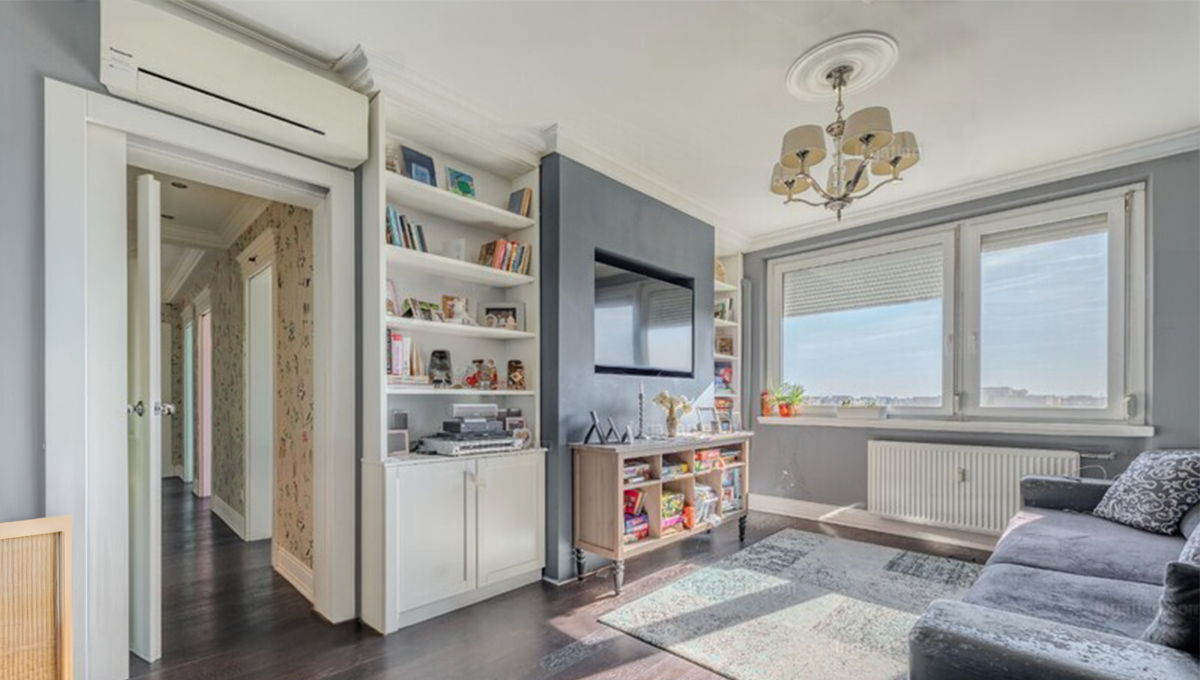 Világos, ízlésesen berendezett nappali nagy ablakokkal és városi kilátással. A falon modern TV, mellette beépített könyvespolc és szekrény található. A helyiséget elegáns csillár, szürke kanapé, dísztárgyak és növények teszik otthonossá, a falakat szürke és fehér árnyalatok uralják.