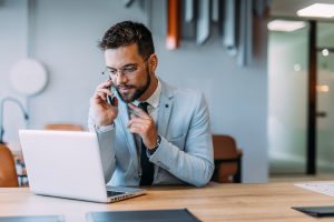 Ingatlanközvetítő öltönyben ül a laptopja előtt, és telefonál.