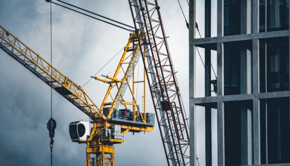 Építkezés részlete: sárga toronydaru és acélszerkezetű épület váza borús égbolt előtt. A kép a modern városi fejlesztéseket és az építőipar tevékenységét ábrázolja, a háttérben fémgerendák és daruk keresztezik egymást.