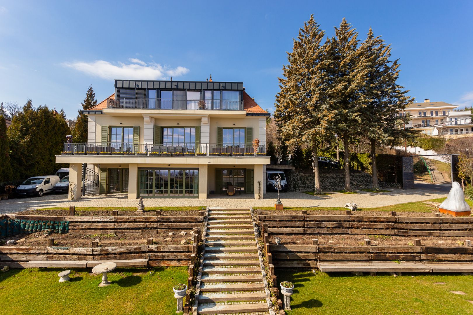 Nagy méretű, elegáns, többszintes villa látható kerttel, enyhén lejtős telken. Az épület világos krémszínű homlokzattal, zöld zsalugáteres ablakokkal és széles erkélyekkel rendelkezik. A felső szinten modern, üvegfelületekkel határolt tetőteraszos kialakítás látható. A ház előtt szimmetrikusan kialakított lépcsősor vezet fel a fő szinthez, két oldalán teraszos kerttel, fa támfalakkal és gondozott zöldfelülettel. Az alsó szinten nagy üvegajtók és fedett terasz található. A kertben dísztárgyak, kerti lépcsők és fenyőfák láthatók, a háttérben további lakóépületekkel. Az udvarban parkoló járművek is megfigyelhetők. Az időjárás derült, kék ég és napsütéses környezet hangsúlyozza az ingatlan exkluzív, nyugodt hangulatát.