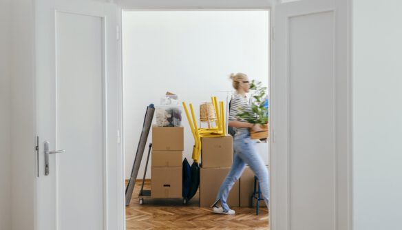 Nyitott ajtókereten keresztül egy világos szobába látni, ahol költözéshez előkészített kartondobozok és felpakolt bútorok állnak. Egy fiatal nő éppen átsétál a helyiségen, kezében növényt tart, miközben a parkettás padlón dobozok, felrakott sárga székek és egy molinóba tekert szőnyeg látható. A kép a költözés vagy új lakásba való beköltözés pillanatát ábrázolja.