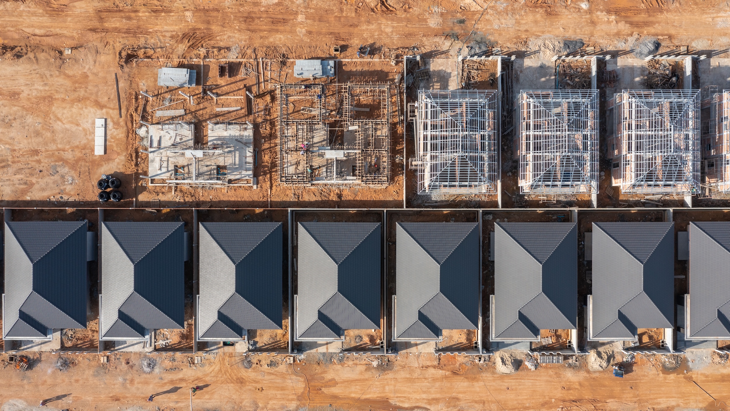 Felülnézeti, drónnal készült felvétel egy lakópark építéséről. Az alsó sorban már elkészült, egyforma tetős családi házak állnak egymás mellett rendezett telkeken, míg a felső részen több ingatlan még különböző építési fázisban van: láthatók alapozások, falazatok és tetőszerkezetek. A terület földes, építési anyagokkal és munkagépekkel, ami aktív kivitelezésre utal.