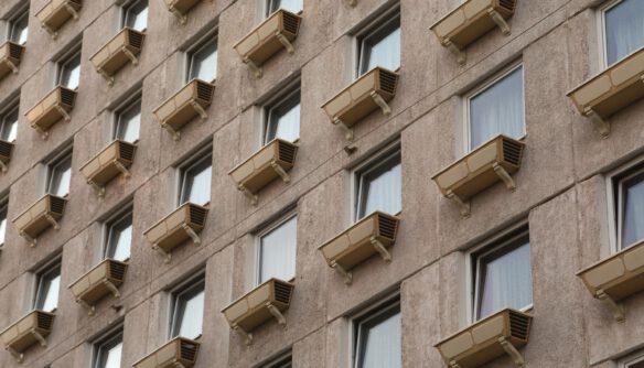 Az épület homlokzatán szabályos rácsszerkezetben elhelyezkedő ablakok láthatók, mindegyik alatt azonos típusú, bézs színű külső légkondicionáló egységgel. A szürke, kissé kopott beton- vagy vakolt felület ipari, paneles jellegre utal. Az ablakok egyformák, fehér keretesek, függönnyel vagy redőnnyel részben takartak. A homlokzat monoton ismétlődése és a klímaberendezések sora sűrűn lakott, többlakásos ház benyomását kelti.
