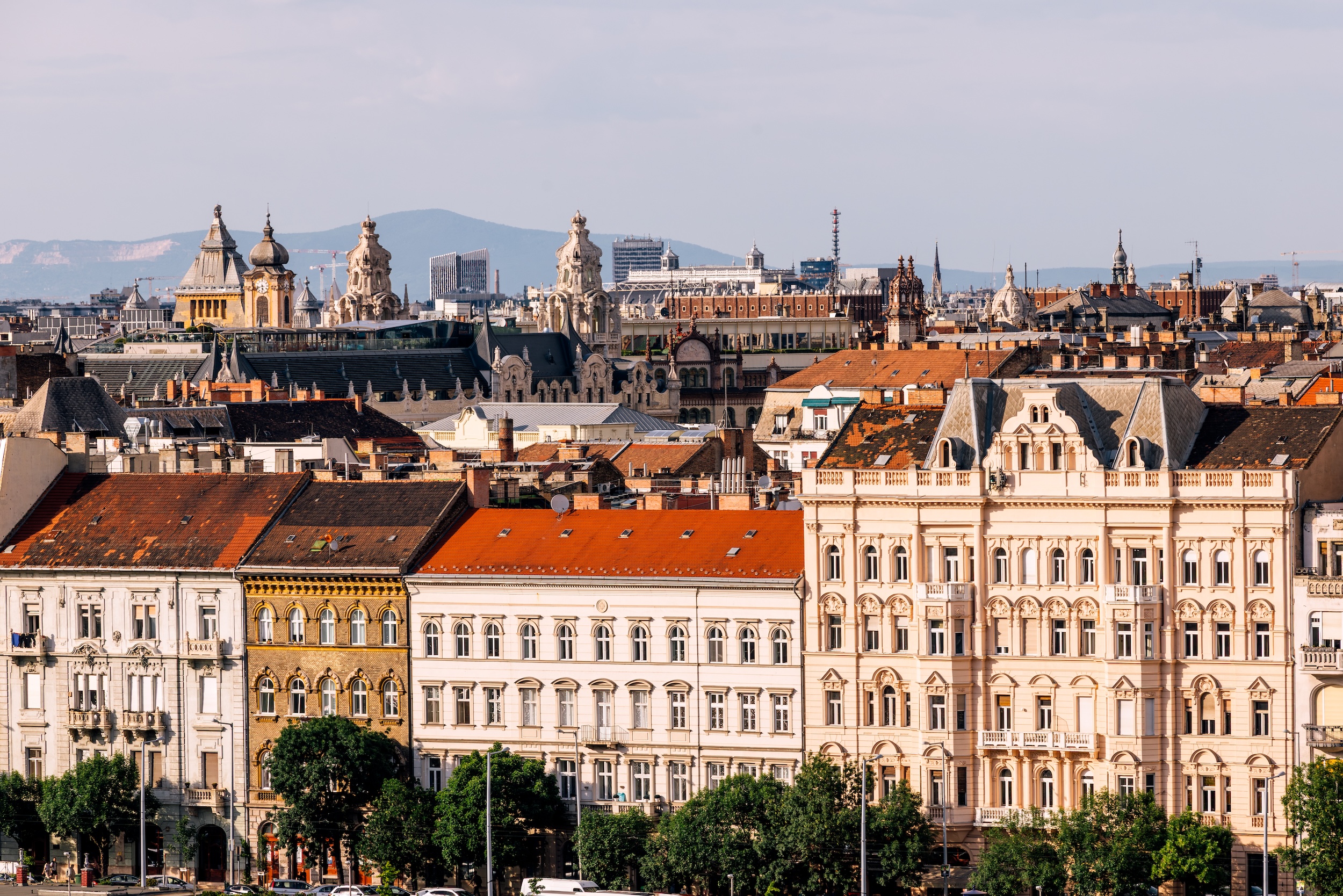 Budapest belvárosának panorámája historikus bérházakkal és díszes homlokzatú épületekkel. Az előtérben világos színű, többemeletes, klasszicista és eklektikus stílusú házak sorakoznak vörös cseréptetőkkel. A háttérben templomtornyok, kupolák és egyéb jellegzetes budapesti épületek emelkednek ki a város látképéből, távolabb pedig hegyek láthatók. A kép a főváros sűrűn beépített, történelmi városszövetét mutatja napsütéses időben.