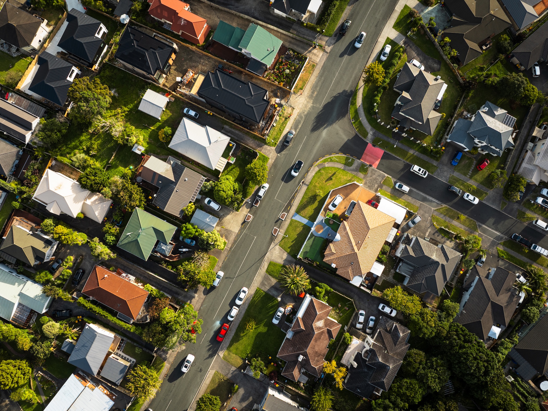 Légi felvétel egy kertvárosi lakóövezetről, ahol családi házak sorakoznak rendezett telkeken. Az utcák sakktáblaszerűen találkoznak egy kereszteződésben, több autó halad és parkol az utak mentén. A házak különböző színű tetőkkel és zöld kertekkel rendelkeznek, a környezetet fák és gondozott udvarok teszik barátságossá.