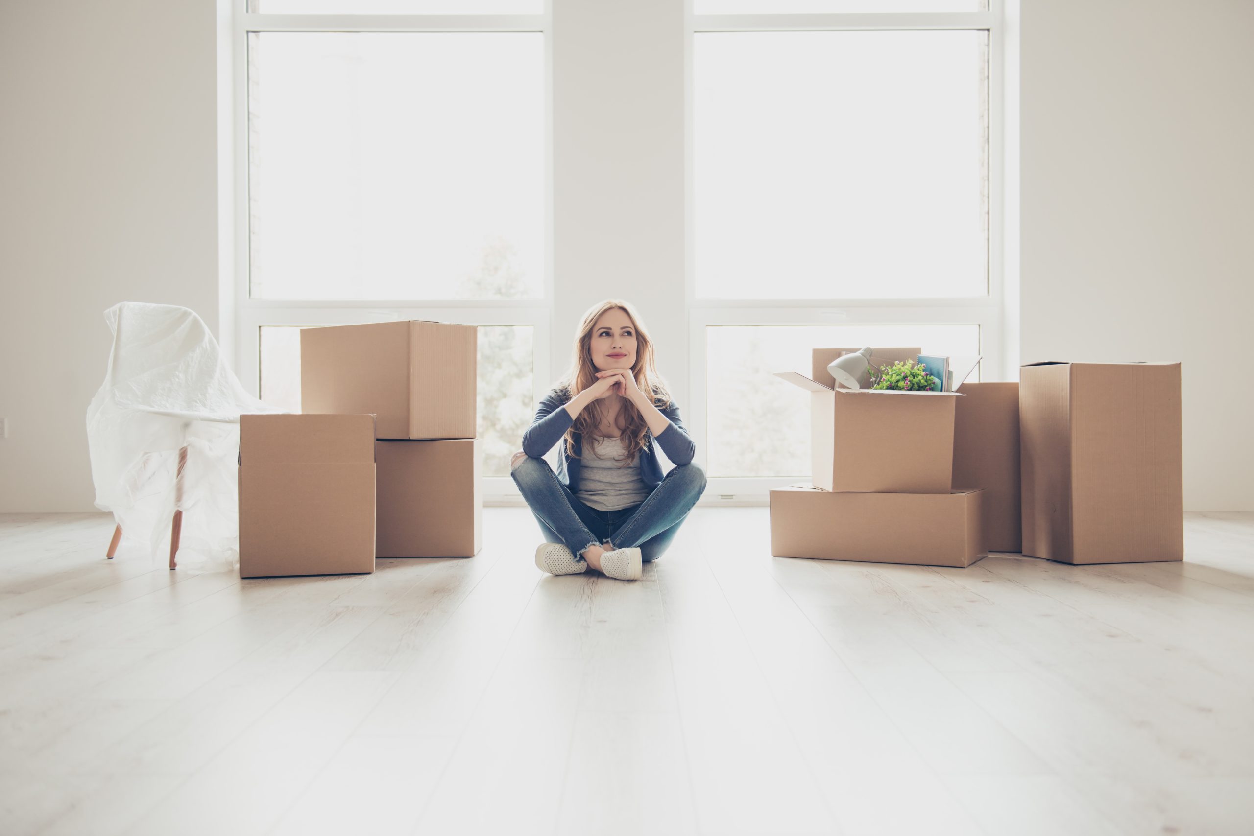 A young woman sits cross-legged on a light wooden floor in a bright, empty apartment. Around her are several cardboard moving boxes, some closed and some open with household items visible inside. A covered chair stands to the left, suggesting recent moving or packing. Large floor-to-ceiling windows behind her let in abundant natural light. She rests her chin on her hands and looks slightly upward with a thoughtful, hopeful expression, as if contemplating a new beginning in the space.